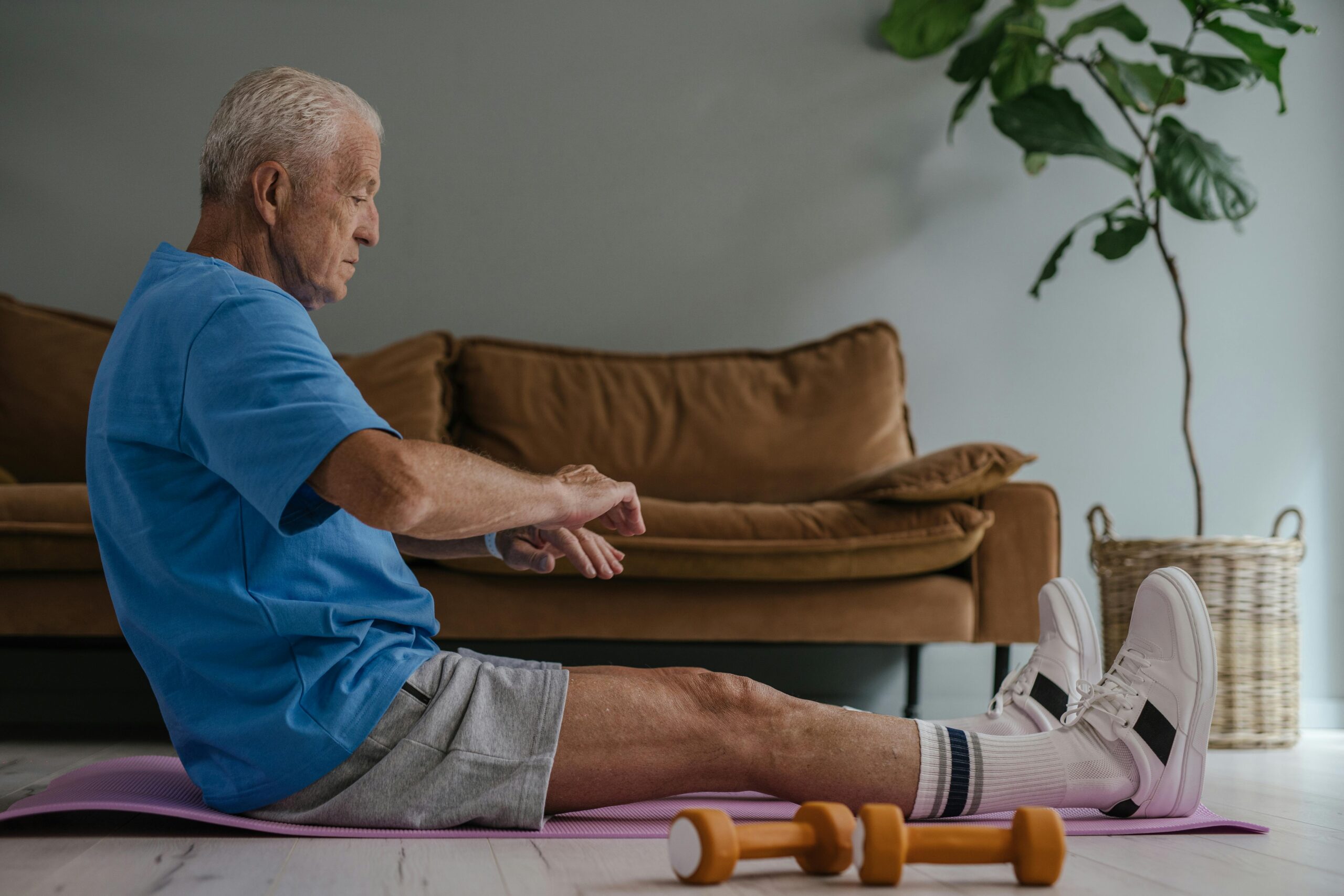 Elderly man in blue shirt exercising on a yoga mat indoors with dumbbells in view.
